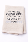 Beige towel with text 'We are the granddaughters of the witches you could not burn' on a white background